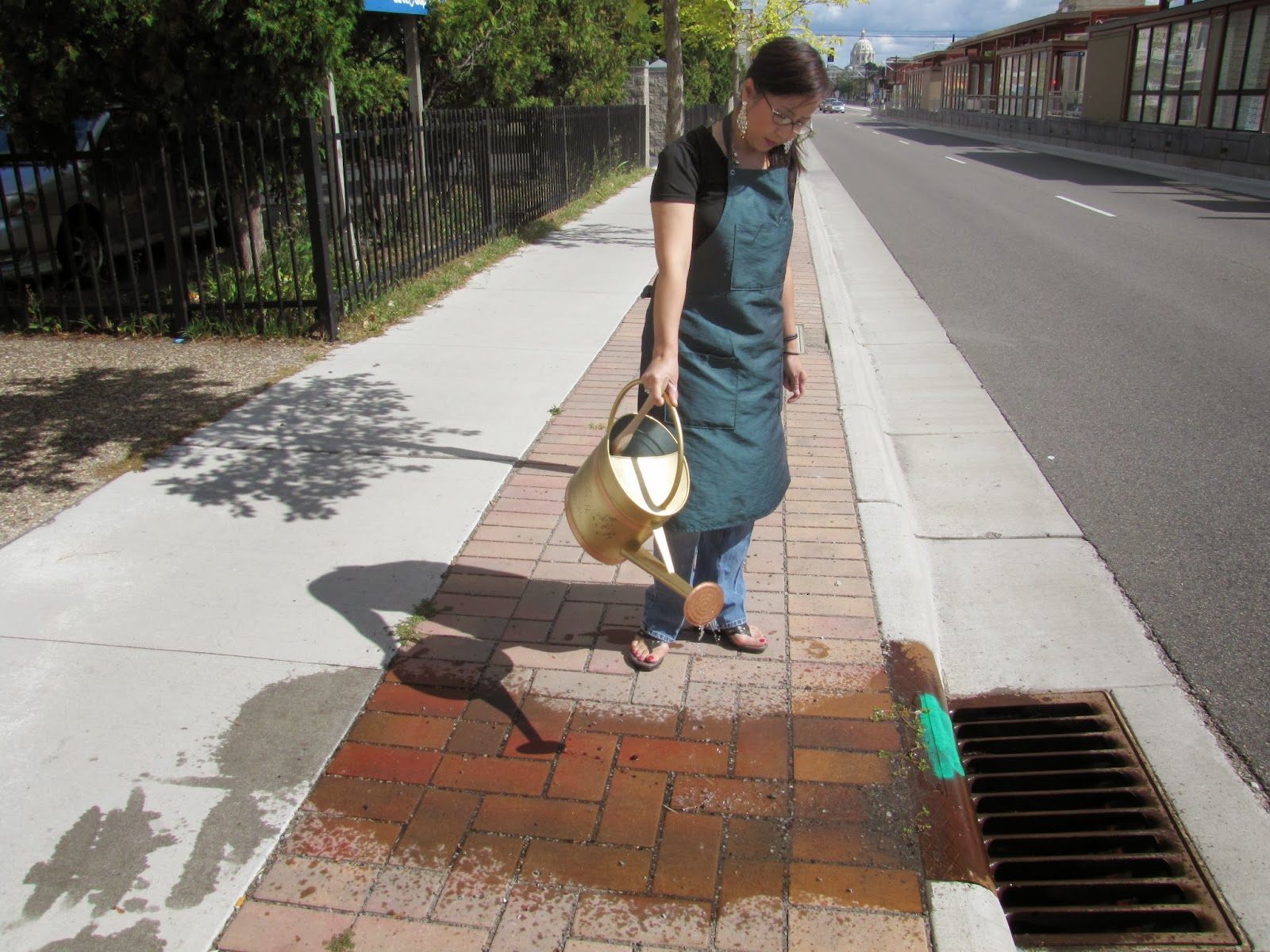 woman in a work apron using a golden watering can to pour water onto the sidewalk near a storm drain